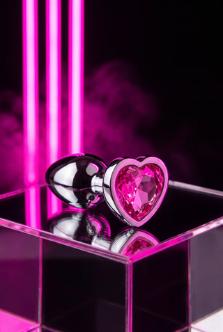 PRETTY SECRET Pink — plug de acero pulido con corazón de cristal rosa sobre podio de acrílico con neón rosa